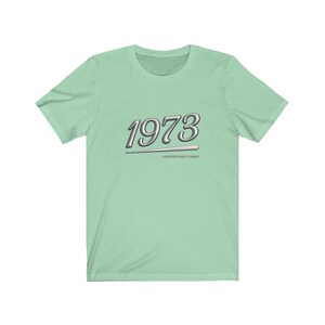 May include: A light green t-shirt with a silver graphic of the year 1973 with a silver line underneath. The text "MADE GREAT IN THE 70'S" is below the year.