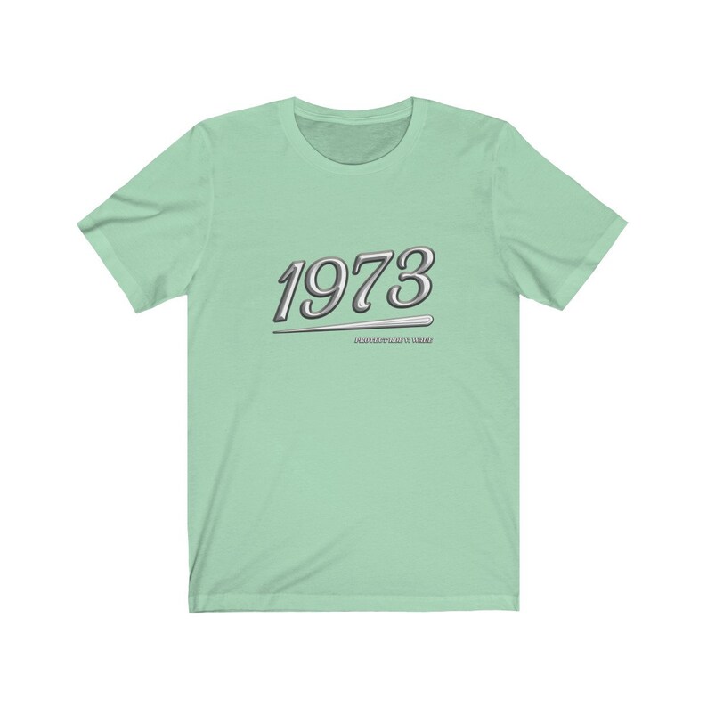 May include: A light green t-shirt with a silver graphic of the year 1973 with a silver line underneath. The text "MADE GREAT IN THE 70'S" is below the year.