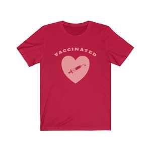 May include: Red short-sleeved t-shirt with the text "VACCINATED" in pink letters and a pink heart with a pink syringe inside.