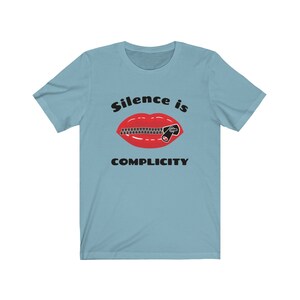 Puede incluir: Una camiseta azul claro con un gr&aacute;fico de una boca roja con una cremallera subida sobre ella. El texto "Silence is Complicity" est&aacute; impreso debajo del gr&aacute;fico.