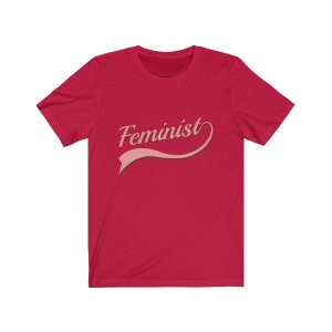 Puede incluir: Camiseta roja con la palabra "Feminista" en letra rosa con una cinta rosa debajo.
