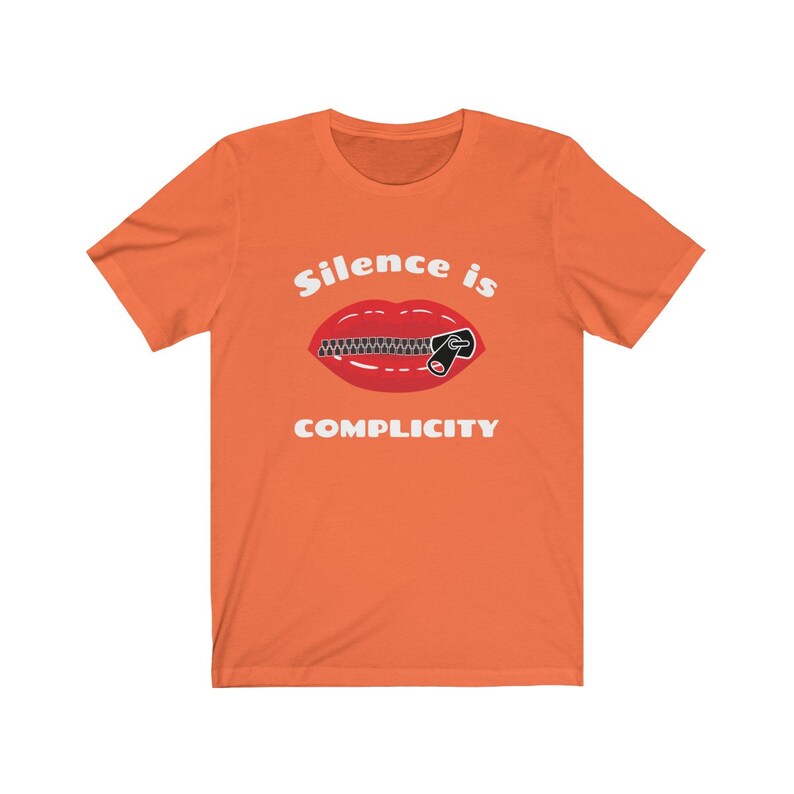 Puede incluir: Camiseta naranja de manga corta con un gr&aacute;fico blanco de una boca roja con una cremallera subida. El texto "Silence is Complicity" est&aacute; impreso debajo del gr&aacute;fico.