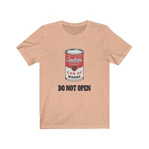 Puede incluir: Una camiseta de color melocot&oacute;n claro con un gr&aacute;fico de una lata roja etiquetada "Caecilians Condensed Can of Worms" con el texto "DO NOT OPEN" debajo de la lata.