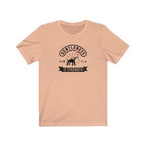 Puede incluir: Camiseta color melocot&oacute;n con un dise&ntilde;o negro. El dise&ntilde;o presenta las palabras "GENTLENESS IS STRENGTH" y una ilustraci&oacute;n de un elefante. El gr&aacute;fico incluye los a&ntilde;os "20" y "21" con estrellas, creando un estilo vintage.