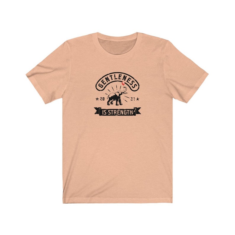 Puede incluir: Camiseta color melocot&oacute;n con un dise&ntilde;o negro. El dise&ntilde;o presenta las palabras "GENTLENESS IS STRENGTH" y una ilustraci&oacute;n de un elefante. El gr&aacute;fico incluye los a&ntilde;os "20" y "21" con estrellas, creando un estilo vintage.