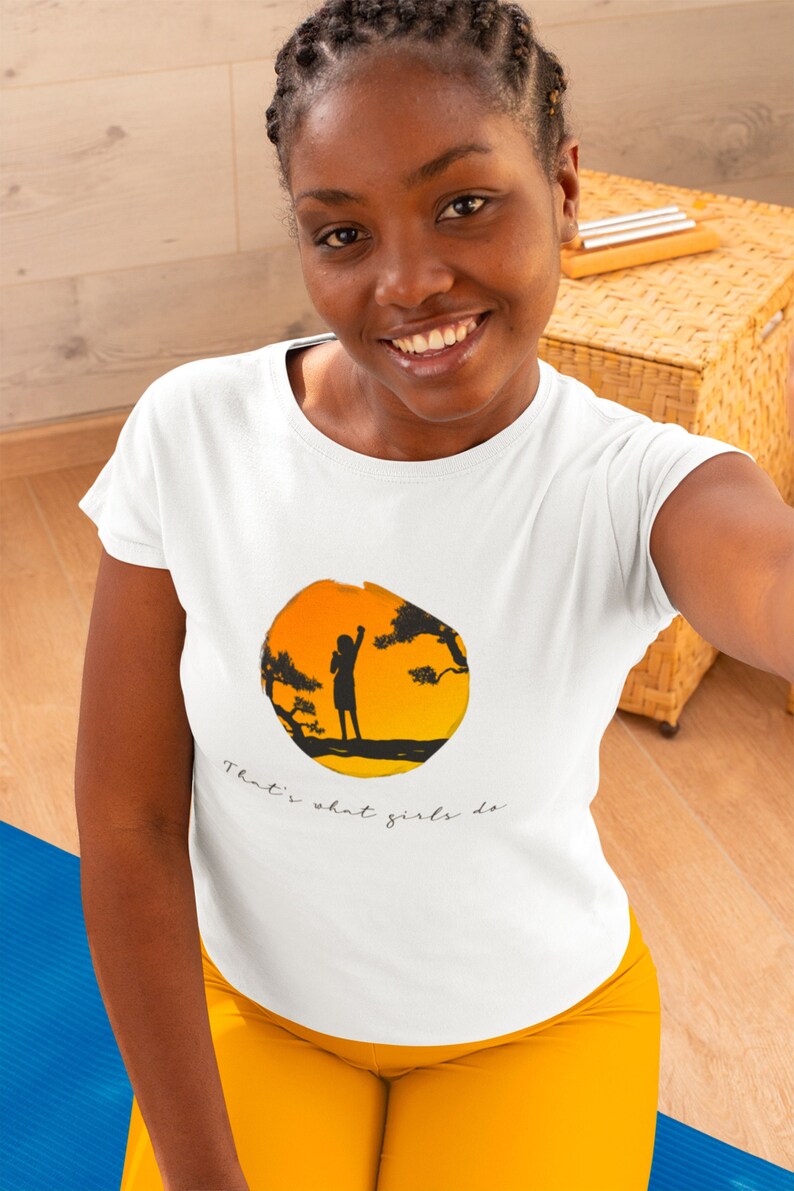 Puede incluir: Camiseta blanca con un gr&aacute;fico de una silueta de una persona de pie en una rama con una puesta de sol detr&aacute;s de ella. El texto "That's what girls do" est&aacute; impreso debajo del gr&aacute;fico.