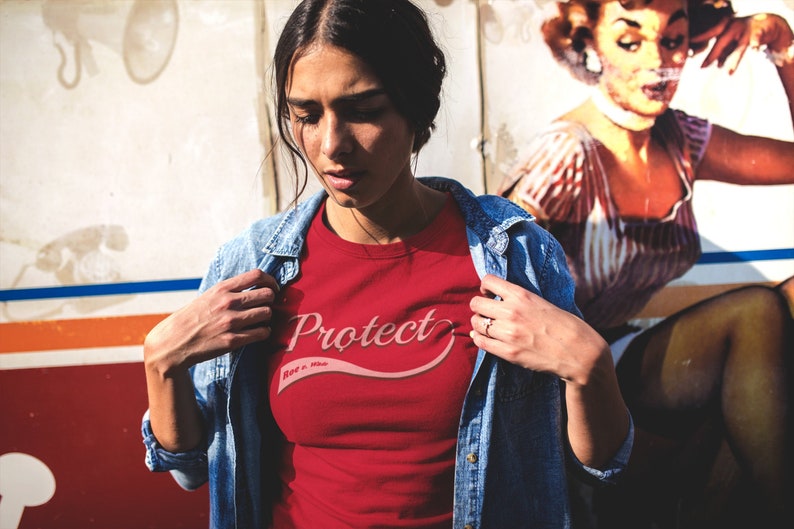 May include: A woman wearing a red t-shirt with the text "Protect More Than This" in a pink cursive font. She is wearing a blue denim jacket over the shirt.
