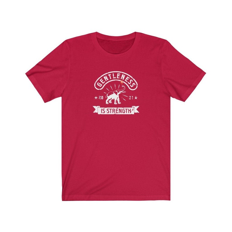 Puede incluir: Una camiseta roja con un gr&aacute;fico blanco. El gr&aacute;fico incluye la palabra "GENTLENESS" en un dise&ntilde;o arqueado sobre una ilustraci&oacute;n de un elefante. Debajo del elefante est&aacute; la frase "IS STRENGTH" en una pancarta. La camiseta tambi&eacute;n tiene los n&uacute;meros "20" y "21" con estrellas.