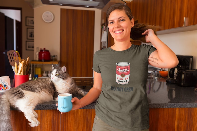 Puede incluir: Una mujer con una camiseta verde con un gr&aacute;fico de una lata de gusanos roja y blanca que dice "Greenthans Can of Worms Do Not Open".