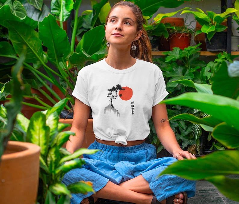 Puede incluir: Una camiseta blanca con una ilustraci&oacute;n en blanco y negro de un &aacute;rbol y un sol rojo. El texto japon&eacute;s en la camiseta dice "呼吸する".