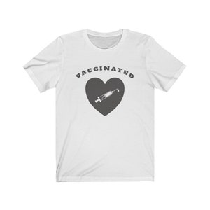 May include: White t-shirt with the text "VACCINATED" in black letters above a gray heart with a white syringe inside.