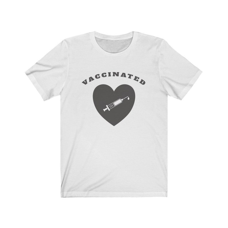 May include: White t-shirt with the text "VACCINATED" in black letters above a gray heart with a white syringe inside.