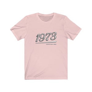 May include: Pink t-shirt with a silver graphic of the year 1973 and the text "Masterfully Crafted Since 1973".