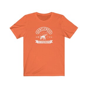 Puede incluir: Camiseta naranja con un gr&aacute;fico blanco. El gr&aacute;fico incluye la palabra "GENTLENESS" en arco, una ilustraci&oacute;n de elefante y la frase "IS STRENGTH" en una pancarta.