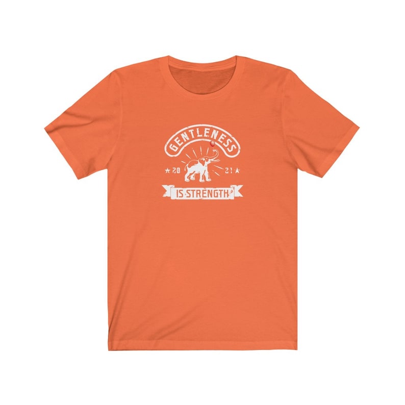 Puede incluir: Camiseta naranja con un gr&aacute;fico blanco. El gr&aacute;fico incluye la palabra "GENTLENESS" en arco, una ilustraci&oacute;n de elefante y la frase "IS STRENGTH" en una pancarta.