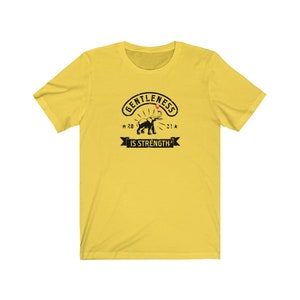 Puede incluir: Camiseta amarilla con un gr&aacute;fico negro que presenta un elefante y el texto "GENTLENESS IS STRENGTH" en un dise&ntilde;o circular. El dise&ntilde;o incluye los a&ntilde;os "20" y "21" con acentos de estrella.