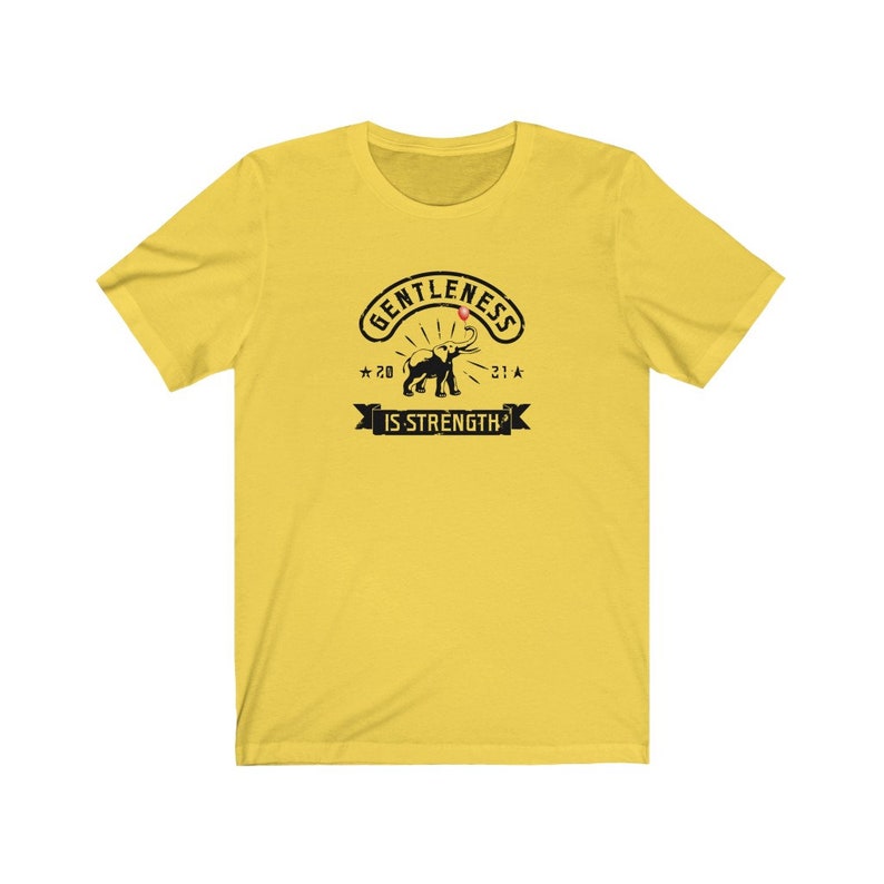 Puede incluir: Camiseta amarilla con un gr&aacute;fico negro que presenta un elefante y el texto "GENTLENESS IS STRENGTH" en un dise&ntilde;o circular. El dise&ntilde;o incluye los a&ntilde;os "20" y "21" con acentos de estrella.