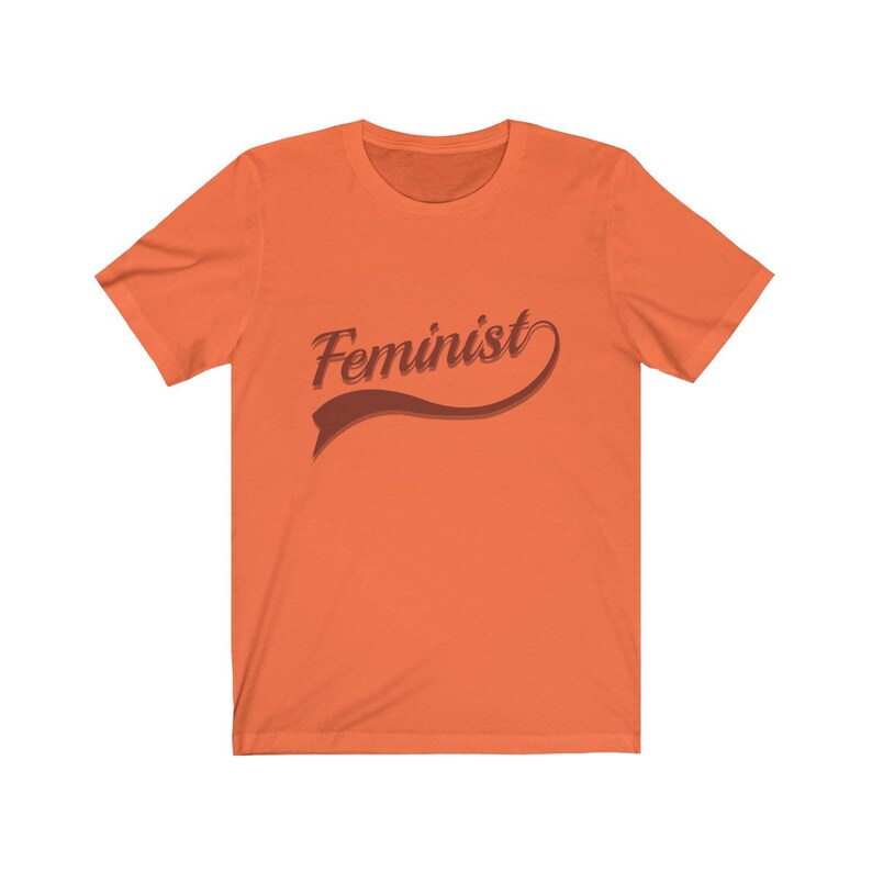 Puede incluir: Camiseta naranja de manga corta con la palabra "Feminista" en una fuente retro marr&oacute;n.