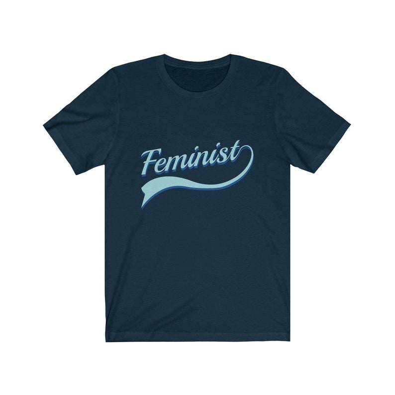 Puede incluir: Una camiseta azul marino con la palabra "Feminista" en una fuente azul claro cursiva con una l&iacute;nea curva azul y blanca detr&aacute;s.