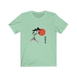 Puede incluir: Una camiseta verde claro con una ilustraci&oacute;n en blanco y negro de un &aacute;rbol en una roca con un sol rojo detr&aacute;s. El texto japon&eacute;s "呼吸する" est&aacute; impreso debajo del &aacute;rbol.
