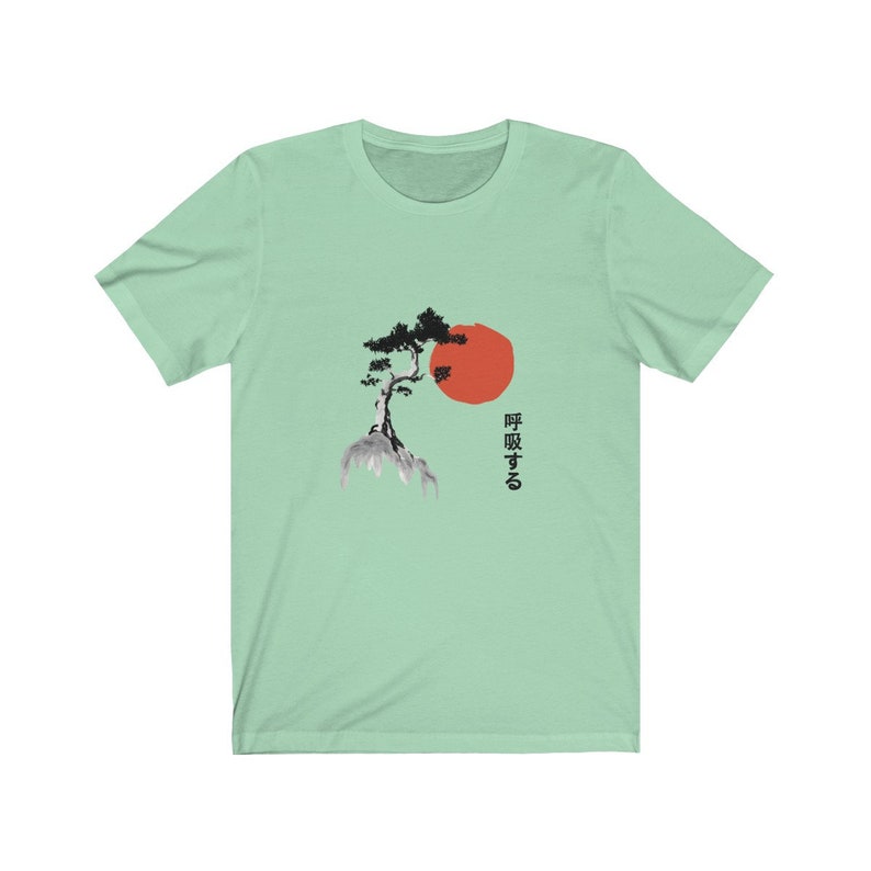 Puede incluir: Una camiseta verde claro con una ilustraci&oacute;n en blanco y negro de un &aacute;rbol en una roca con un sol rojo detr&aacute;s. El texto japon&eacute;s "呼吸する" est&aacute; impreso debajo del &aacute;rbol.