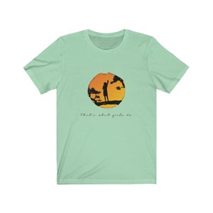 Puede incluir: Una camiseta verde claro con la silueta de una persona de pie en una rama de un &aacute;rbol frente a una puesta de sol. El texto "That's what girls do" est&aacute; impreso debajo de la imagen.