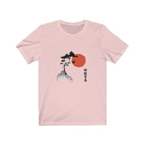 Puede incluir: Camiseta rosa con una ilustraci&oacute;n en blanco y negro de un &aacute;rbol en una monta&ntilde;a con un sol rojo detr&aacute;s. El texto japon&eacute;s "呼吸する" est&aacute; impreso en el lado derecho de la camiseta.