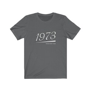 May include: A dark gray t-shirt with the year 1973 printed in white with a silver underline. The text "PROTECT ROE V. WADE" is printed in white below the year.