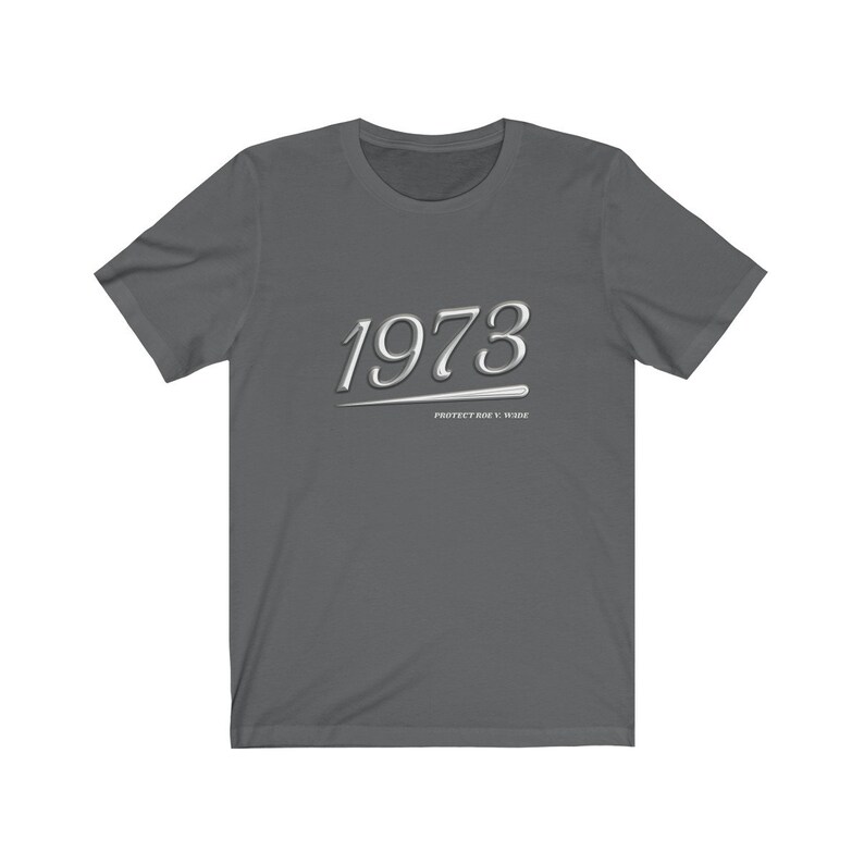 May include: A dark gray t-shirt with the year 1973 printed in white with a silver underline. The text "PROTECT ROE V. WADE" is printed in white below the year.