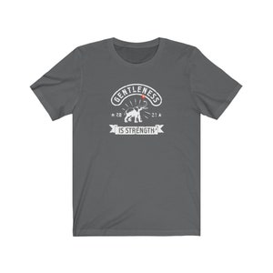 Puede incluir: Camiseta gris oscuro con un gr&aacute;fico blanco. El gr&aacute;fico presenta la palabra "GENTLENESS" en arco, un elefante y las palabras "IS STRENGTH" en una pancarta. La camiseta tiene mangas cortas y cuello redondo.