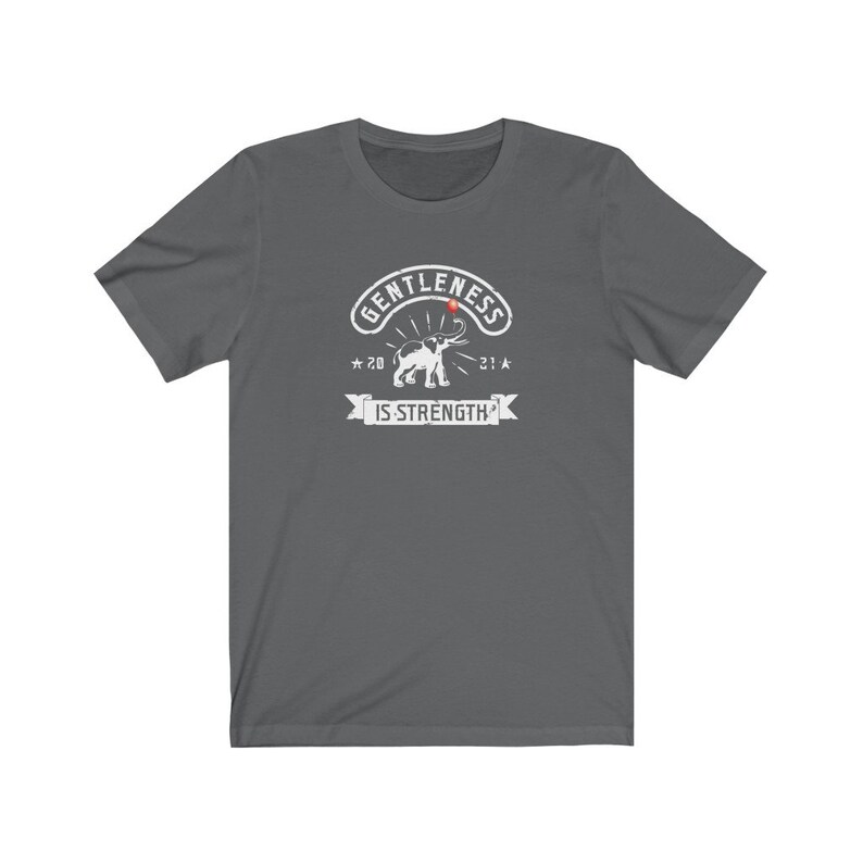 Puede incluir: Camiseta gris oscuro con un gr&aacute;fico blanco. El gr&aacute;fico presenta la palabra "GENTLENESS" en arco, un elefante y las palabras "IS STRENGTH" en una pancarta. La camiseta tiene mangas cortas y cuello redondo.