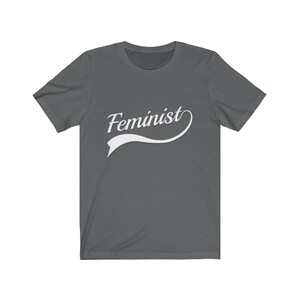 Puede incluir: Una camiseta gris oscuro con un dise&ntilde;o curvo blanco que dice "Feminista".