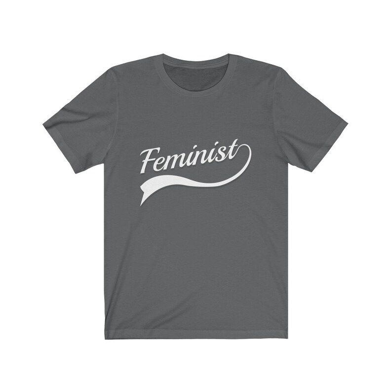 Puede incluir: Una camiseta gris oscuro con un dise&ntilde;o curvo blanco que dice "Feminista".