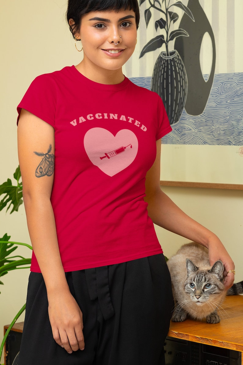 May include: A red t-shirt with a pink heart and a syringe graphic. The text "VACCINATED" is printed in white letters above the heart.