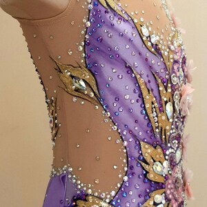 Puede incluir: Un maillot sin mangas con una base de tela morada clara y paneles de malla color carne, adornado con aplicaciones doradas en forma de hoja, numerosos cristales transparentes y morados, y peque&ntilde;os detalles florales rosas en el frente.