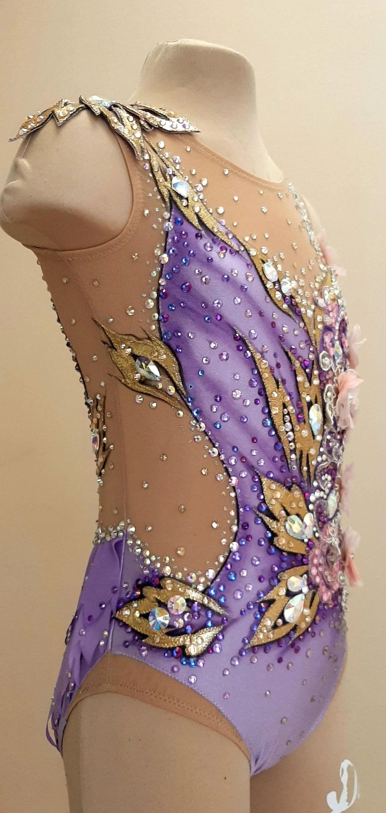 Puede incluir: Un maillot sin mangas con una base de tela morada clara y paneles de malla color carne, adornado con aplicaciones doradas en forma de hoja, numerosos cristales transparentes y morados, y peque&ntilde;os detalles florales rosas en el frente.
