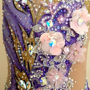 Puede incluir: Un maillot con una base en degradado morado y morado claro con paneles de malla color carne. La prenda est&aacute; decorada con cristales transparentes y morados, detalles de tela dorada y varias flores de tela superpuestas en rosa p&aacute;lido.