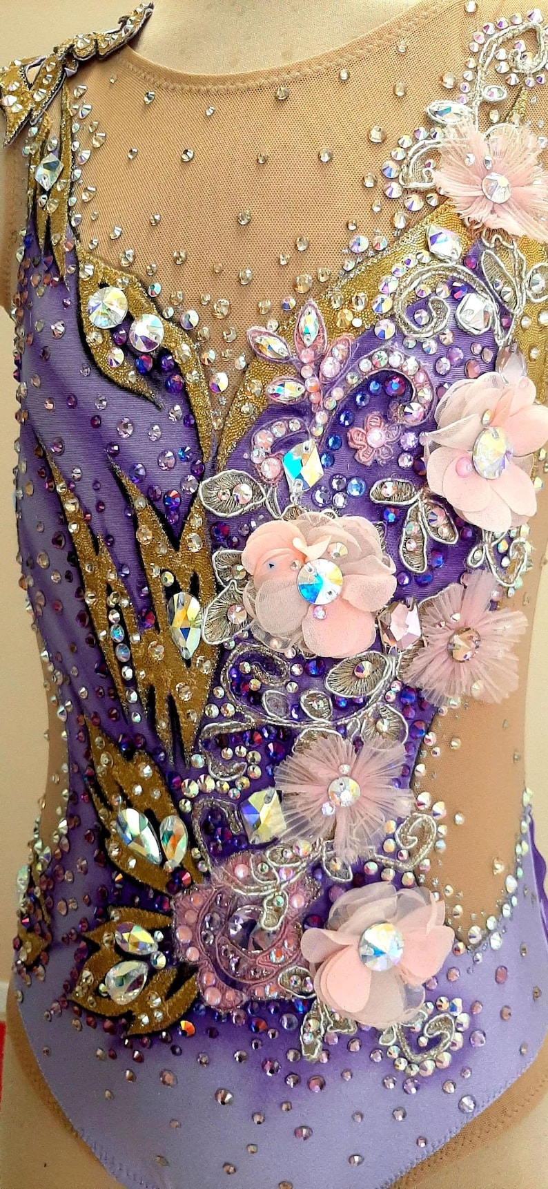 Puede incluir: Un maillot con una base en degradado morado y morado claro con paneles de malla color carne. La prenda est&aacute; decorada con cristales transparentes y morados, detalles de tela dorada y varias flores de tela superpuestas en rosa p&aacute;lido.