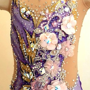 Puede incluir: Un maillot sin mangas con tejido en degradado morado, aplicaciones doradas en forma de hoja, cristales transparentes y flores de tela rosa p&aacute;lido. El cuerpo incluye paneles de malla color carne en el escote y los laterales con pedrer&iacute;a.