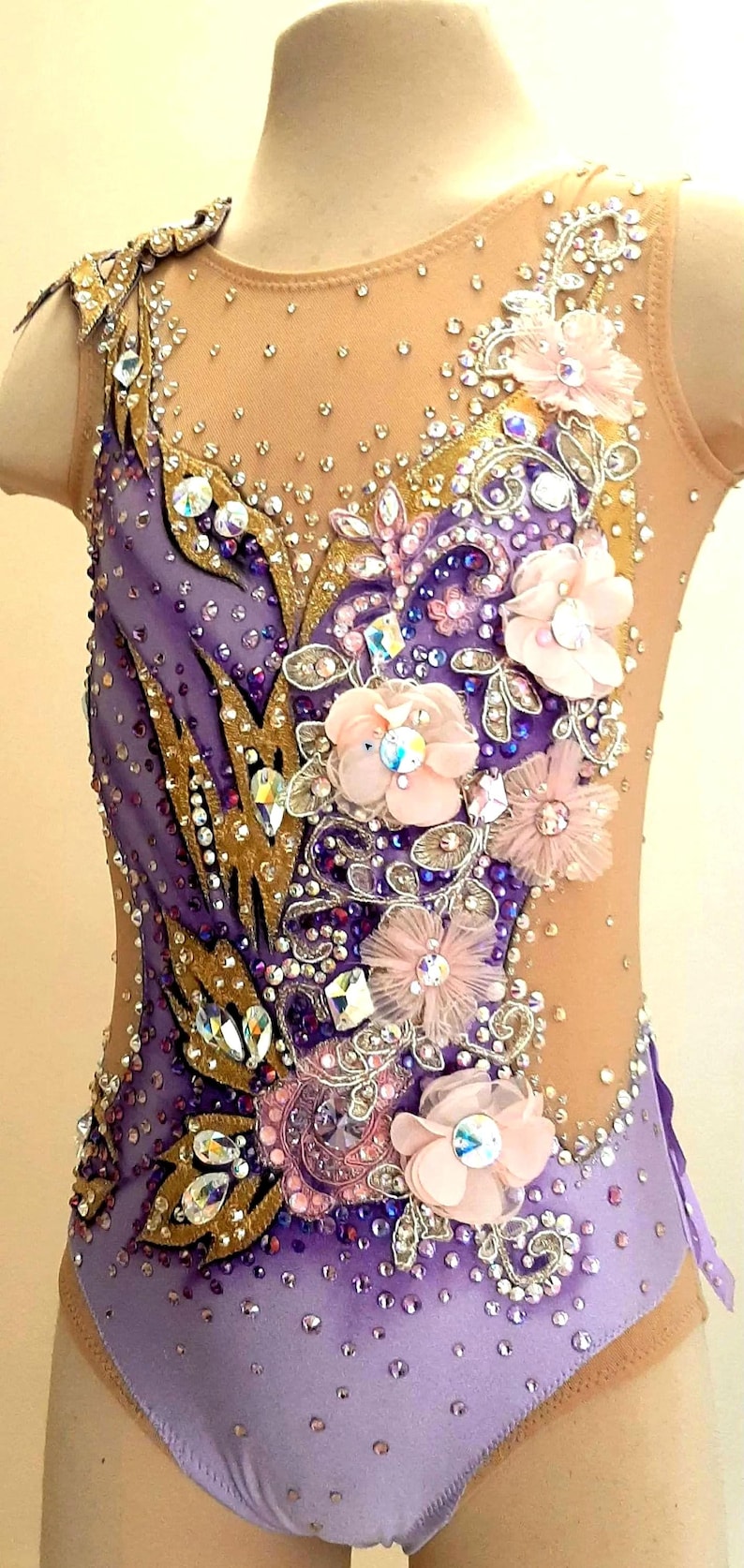 Puede incluir: Un maillot sin mangas con tejido en degradado morado, aplicaciones doradas en forma de hoja, cristales transparentes y flores de tela rosa p&aacute;lido. El cuerpo incluye paneles de malla color carne en el escote y los laterales con pedrer&iacute;a.