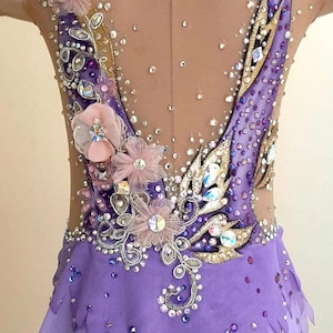 Puede incluir: Un maillot sin mangas con una base de tela morada y una secci&oacute;n superior de malla color carne. La prenda est&aacute; profusamente decorada con cristales transparentes y morados, detalles met&aacute;licos dorados y flores de tela rosa p&aacute;lido.