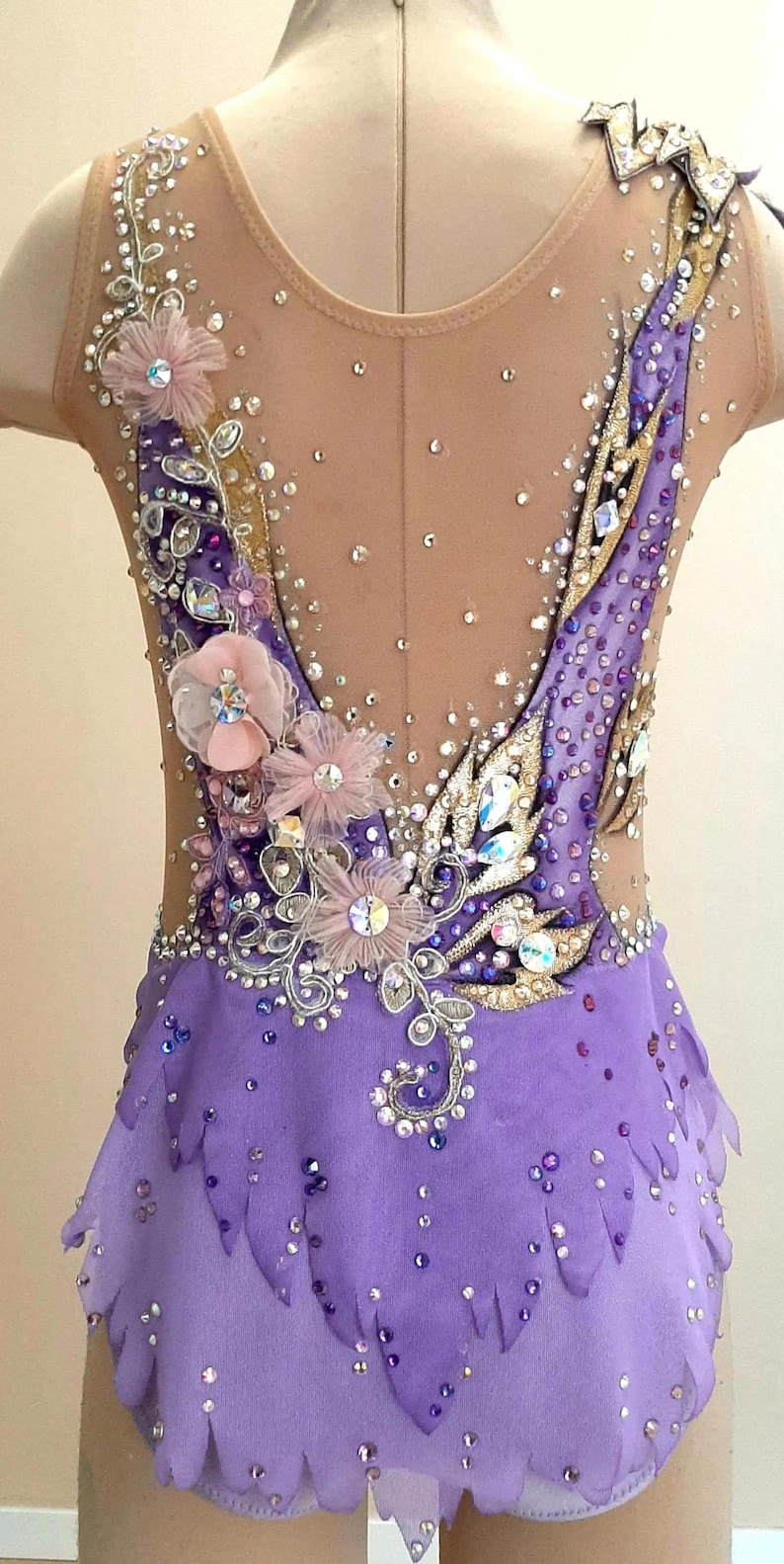 Puede incluir: Un maillot sin mangas con una base de tela morada y una secci&oacute;n superior de malla color carne. La prenda est&aacute; profusamente decorada con cristales transparentes y morados, detalles met&aacute;licos dorados y flores de tela rosa p&aacute;lido.