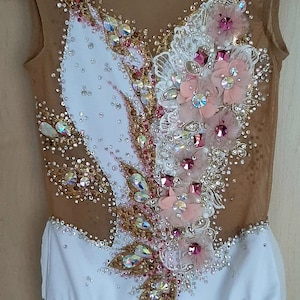 May include: A white leotard with a nude mesh overlay. The leotard is decorated with pink and gold flowers, rhinestones, and sequins.