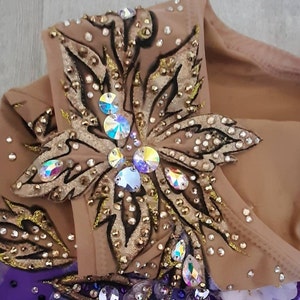 Puede incluir: Un maillot beige con una compleja aplicaci&oacute;n en forma de flor en tonos dorados y bronce, decorado con cristales iridiscentes y peque&ntilde;os remaches met&aacute;licos. El dise&ntilde;o incluye contornos oscuros y una secci&oacute;n de tela morada en la base.