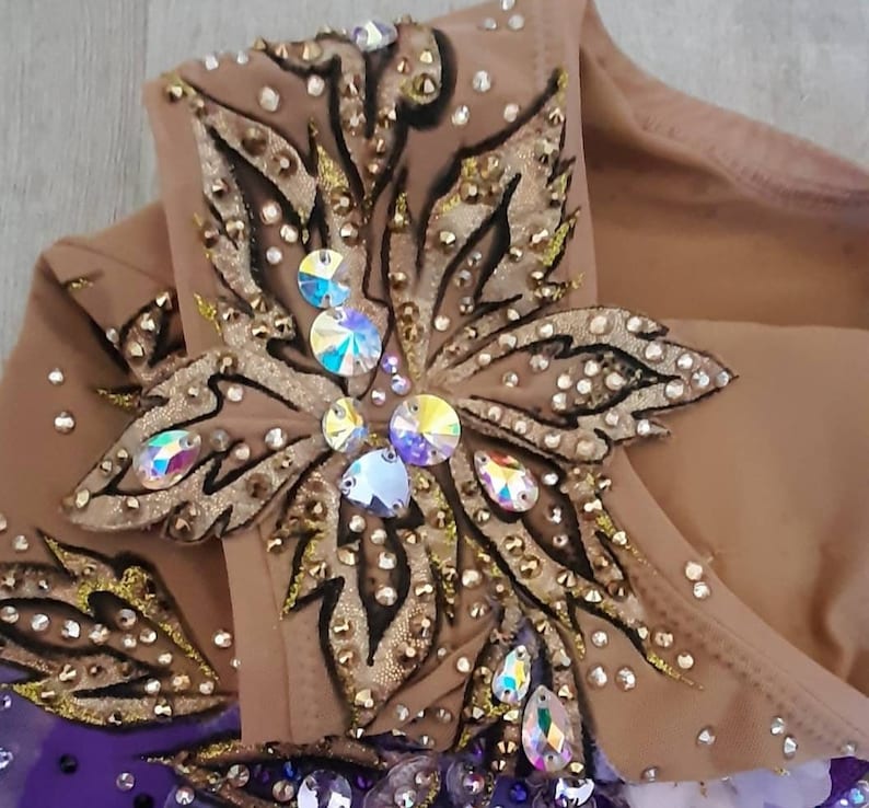 Puede incluir: Un maillot beige con una compleja aplicaci&oacute;n en forma de flor en tonos dorados y bronce, decorado con cristales iridiscentes y peque&ntilde;os remaches met&aacute;licos. El dise&ntilde;o incluye contornos oscuros y una secci&oacute;n de tela morada en la base.