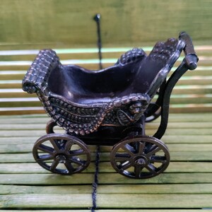 May include: A miniature black metal baby carriage with ornate details and wheels. The carriage is sitting on a bamboo surface.
