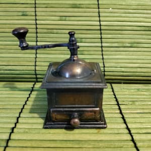 May include: A vintage-style metal pencil sharpener shaped like a coffee grinder. The sharpener is brown and has a crank on the side. It is sitting on a green bamboo mat.