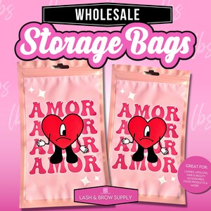 May include: Two pink resealable plastic bags with a white border. Each bag has a cartoon heart with a sad face and the word "AMOR" repeated four times in a pink and white font. The text "LASH & BROW SUPPLY" is printed on the bottom of the bags. The text "GREAT FOR: LASHES, LIPGLOSS, HAIR & BEAUTY ACCESSORIES, FOOD PRODUCTS & MORE!" is printed on the right bag.