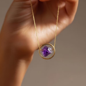 Pode incluir: Um colar dourado com um pendente circular contendo uma gema roxa vibrante. O colar é exibido contra um fundo neutro, destacando o design e a cor da joia.