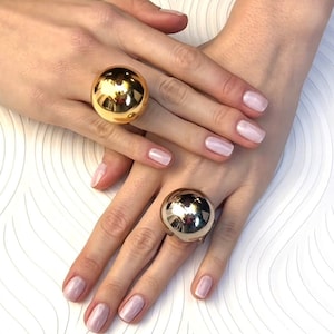 May include: Two gold-toned rings with large, round, smooth, sphere-shaped tops. The rings are worn on the fingers of a person's hands.